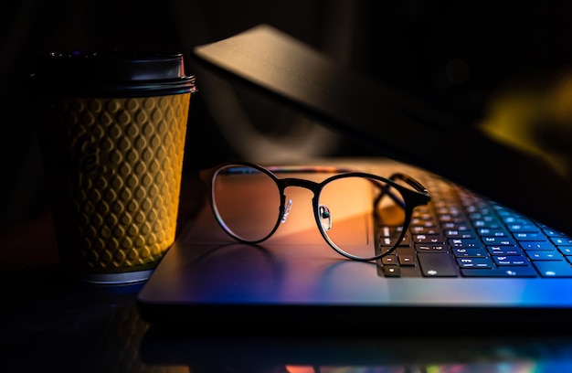 Photo gratuite un ordinateur portable à moitié fermé dans le noir avec des verres lumineux colorés et une tasse en papier