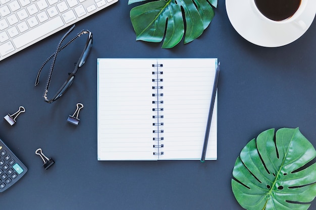 Ordinateur portable près de papeterie, clavier et lunettes sur une table bleu foncé avec pinces pour calculatrice et reliure