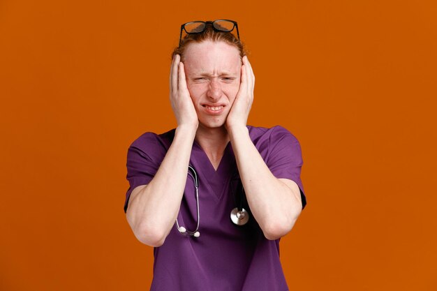 Oreilles couvertes agacées jeune médecin de sexe masculin portant l'uniforme avec stéthoscope isolé sur fond orange