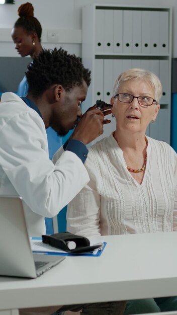 Otologue faisant une consultation d'oreille pour un patient âgé à l'aide d'un otoscope professionnel dans un établissement de santé. Spécialiste de l'outil en acier moderne examinant l'infection alors qu'il était assis au bureau dans l'armoire