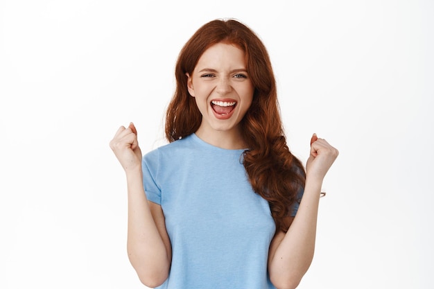 Ouais je l'ai fait. Fille championne aux cheveux rouges célébrant, gagnant le prix, criant de joie et de satisfaction de la victoire, triomphant, debout sur fond blanc