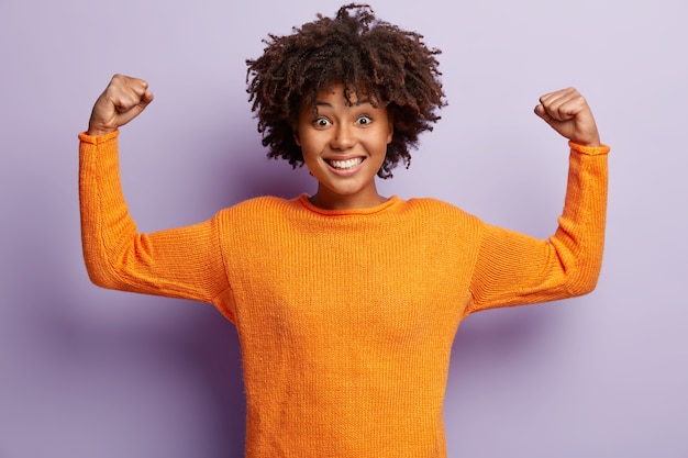 Photo gratuite oui, je suis gagnant! joyeuse femme souriante à la peau sombre lève les bras, démontre les muscles, étant puissante