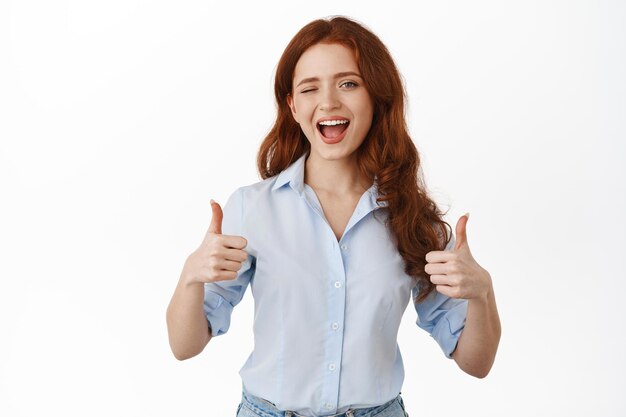 Oui très bien. Femme rousse souriante acclamant, faisant un clin d'œil et montrant les pouces vers le haut en signe d'approbation, louant un excellent travail, un bon travail, faisant un compliment, debout en blouse sur fond blanc