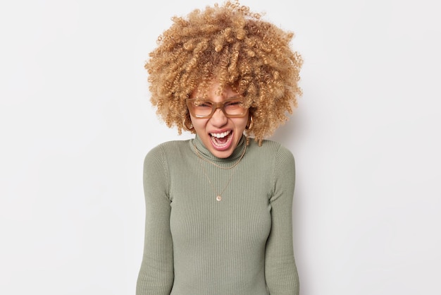 Photo gratuite outragée émotionnelle jeune femme bouclée crie avec colère exprime sa rage déteste quelque chose querelle avec quelqu'un porte un col roulé et des lunettes isolées sur fond blanc. émotions négatives