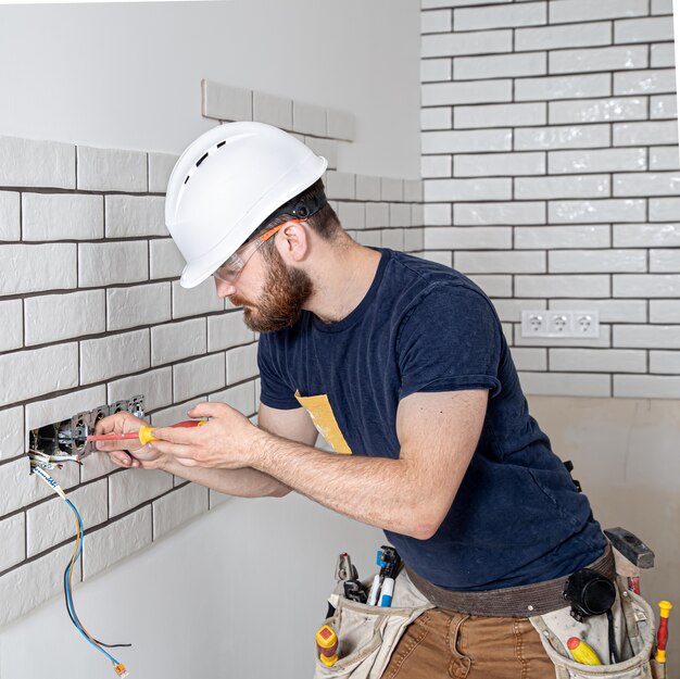 Ouvrier électricien avec une barbe en salopette lors de l'installation de prises. Concept de rénovation domiciliaire.