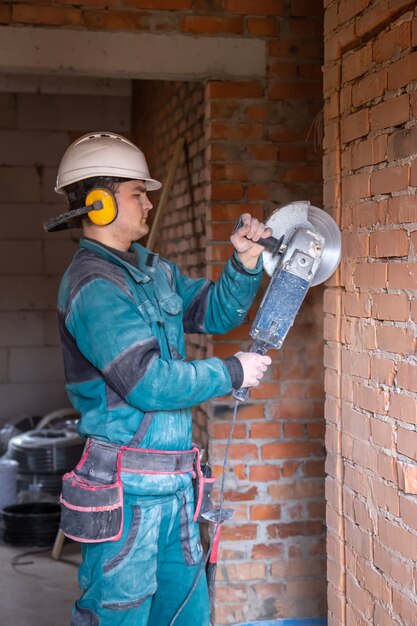Un ouvrier électricien dans un casque de protection dans un lieu de travail travaille avec une meuleuse.