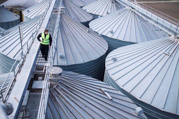 Ouvrier d'usine marchant sur une plate-forme métallique et faisant une inspection visuelle sur des réservoirs ou des silos de stockage d'aliments industriels