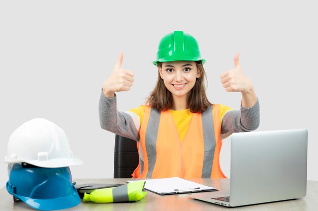 Ouvrière en uniforme assise au bureau et montrant les pouces vers le haut. Photo de haute qualité