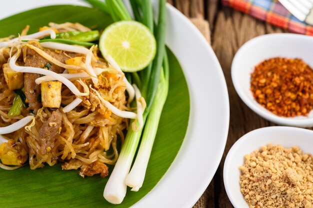 Pad Thai dans une assiette blanche avec du citron, des œufs et des assaisonnements sur une table en bois.