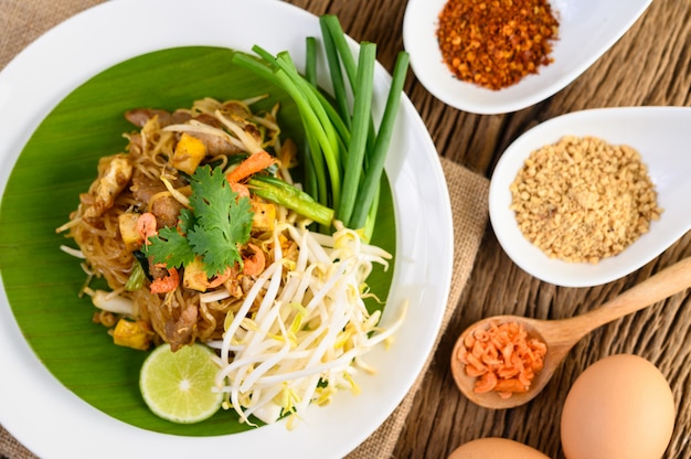 Pad Thai dans une assiette blanche avec du citron, des œufs et des assaisonnements sur une table en bois.