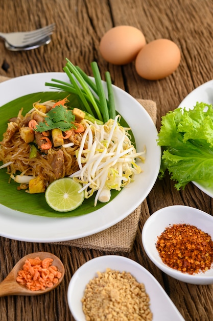 Pad Thai dans une assiette blanche avec du citron, des œufs et des assaisonnements sur une table en bois.