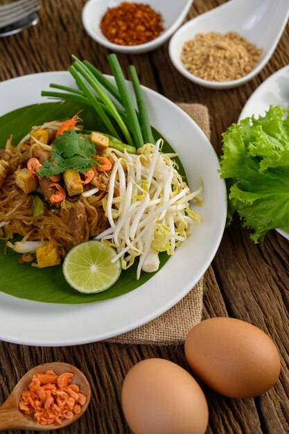 Pad Thai dans une assiette blanche avec du citron, des œufs et des assaisonnements sur une table en bois.