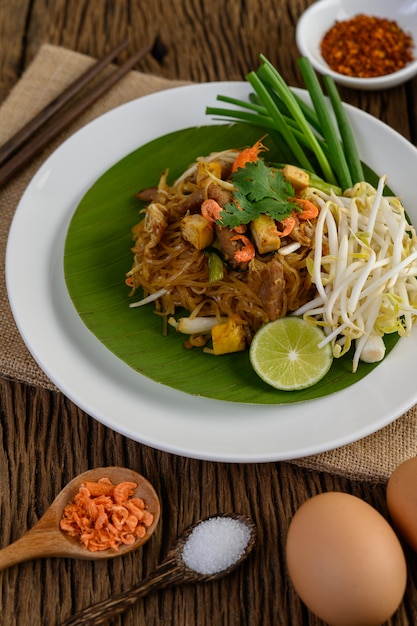 Pad Thai dans une assiette blanche avec du citron, des œufs et des assaisonnements sur une table en bois.