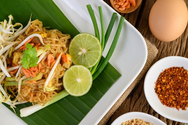 Pad Thai dans une assiette blanche avec du citron, des œufs et des assaisonnements sur une table en bois.