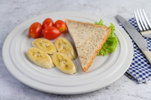 Pain, banane et tomate sur une plaque blanche avec une fourchette et un couteau.