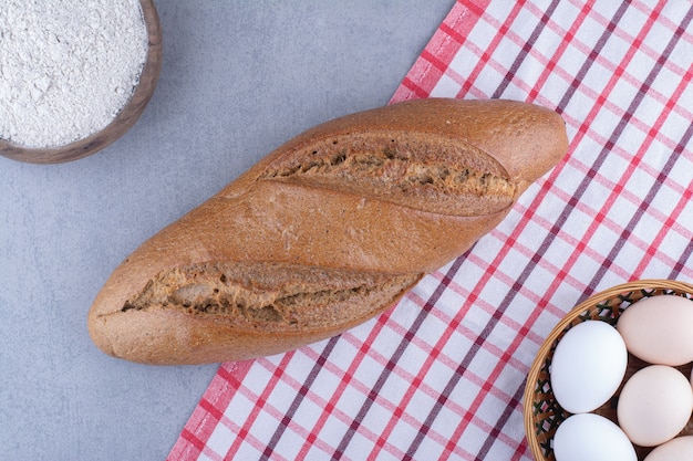 Pain bâton à côté d'un panier d'oeufs et un bol de farine sur une surface en marbre