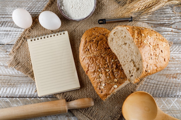 Pain coupé en deux avec un bloc-notes, des œufs, un rouleau à pâtisserie sur un tissu de sac et une surface en bois, vue de dessus.