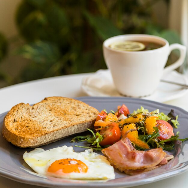 Pain grillé; œuf à moitié frit; salade et bacon sur une assiette grise près de la tasse de thé