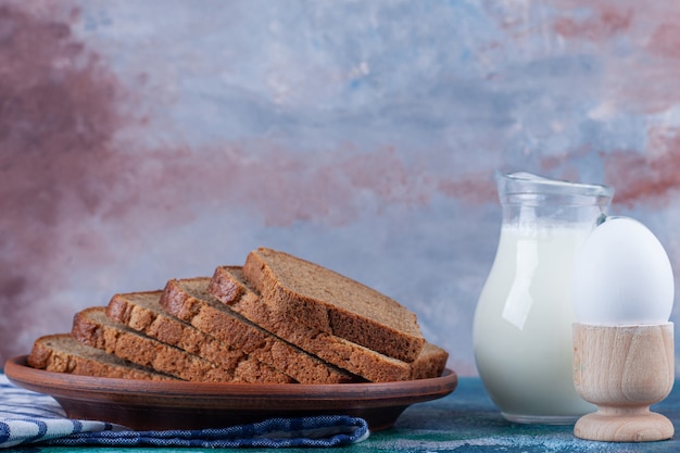 Photo gratuite pain de seigle sur une assiette à côté d'une cruche de lait, farine et œuf sur une serviette sur bleu.