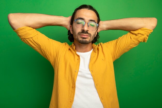 Paisible jeune bel homme caucasien portant des lunettes en gardant les mains derrière la tête avec les yeux fermés isolé sur fond vert