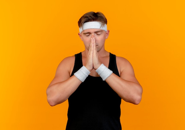 Paisible jeune bel homme sportif portant bandeau et bracelets mettant les mains dans le geste de prier près du visage avec les yeux fermés isolé sur fond orange