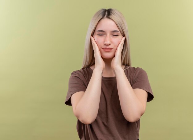 Paisible jeune fille blonde mettant les mains sur les joues avec les yeux fermés sur un espace vert isolé avec copie espace