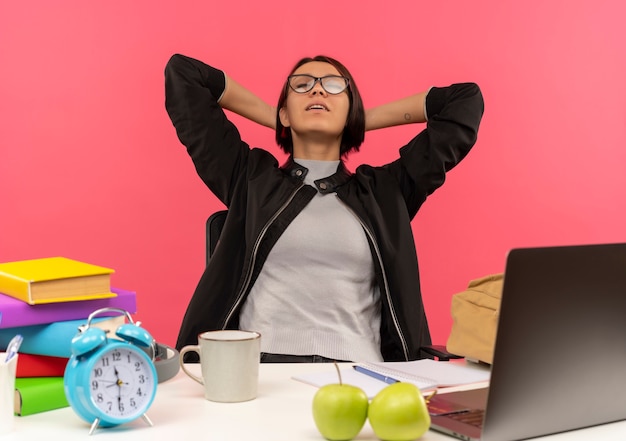 Paisible jeune fille étudiante portant des lunettes assis au bureau en mettant les mains derrière le cou avec les yeux fermés isolé sur fond rose