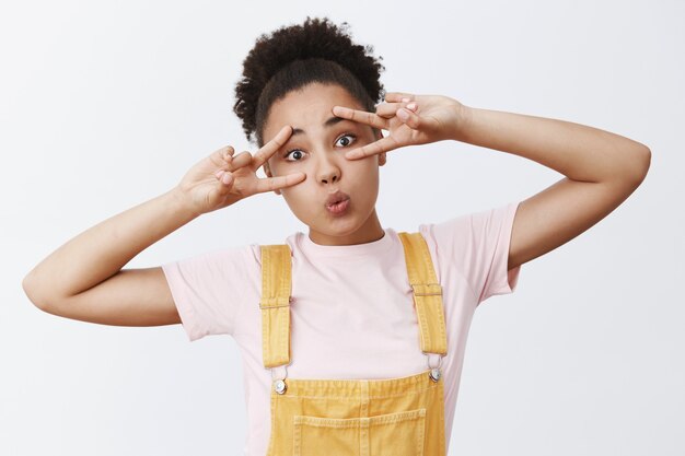 Paix dans nos cœurs. Jolie fille afro-américaine moderne à la mode en salopette jaune sur t-shirt, tête inclinable, pliant les lèvres en baiser et montrant des signes de victoire sur les yeux joyeusement, dans une ambiance disco