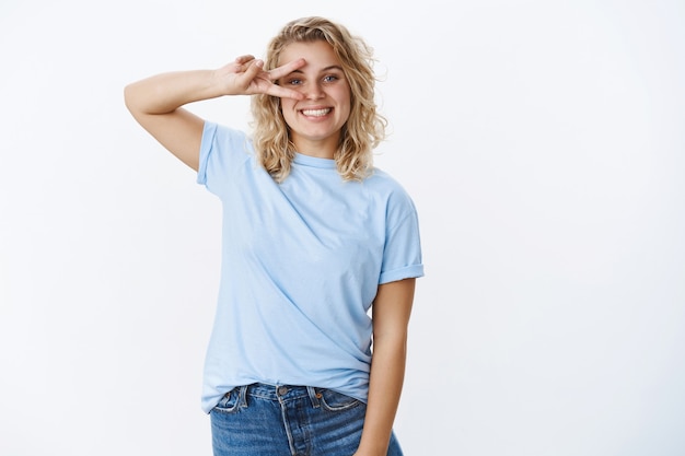 Photo gratuite paix mon ami. portrait d'une étudiante européenne mignonne, heureuse et gentille aux yeux bleus et aux cheveux courts et bouclés montrant la victoire ou le geste disco sur les yeux et souriante positive, de bonne humeur