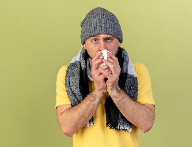 Pâle jeune homme slave malade blonde portant chapeau d'hiver et écharpe boit tasse de thé isolé sur mur vert olive avec espace copie