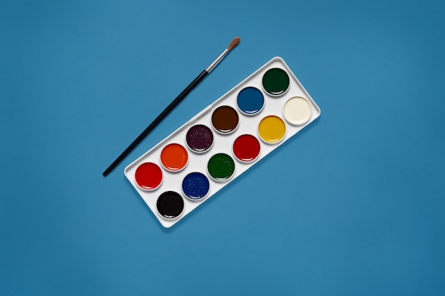 Photo gratuite palette blanche avec douze couleurs différentes au centre de l'image, sans con, pinceau noir à côté des peintures. couleur bleu fantôme. concept d'art. équipement nécessaire pour la peinture.