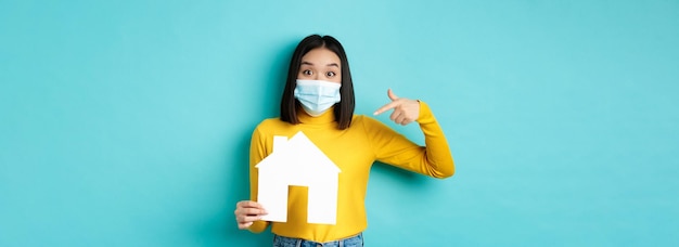 Photo gratuite pandémie de covid et concept immobilier joyeuse femme asiatique souriante dans un masque médical montrant du papier ho