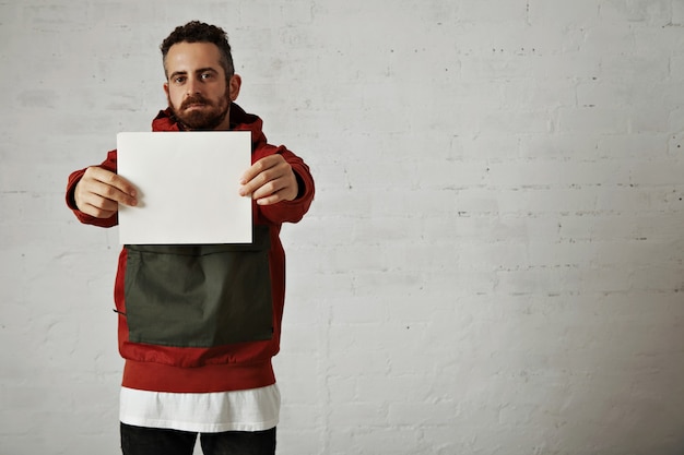 Panneau blanc uni tenu par un beau modèle masculin attrayant avec barbe portant une veste rouge et grise, un jean et un t-shirt blanc isolé sur blanc