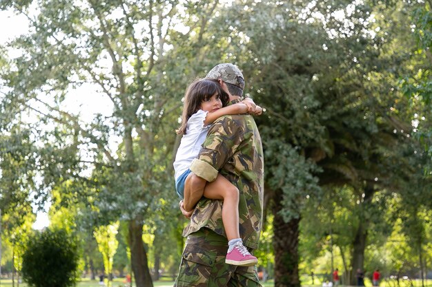 Papa caucasien en uniforme de l'armée embrassant sa fille. Père d'âge moyen debout dans le parc de la ville. Jolie fille assise sur ses mains et étreignant papa sur le cou. Enfance, week-end et concept de parent militaire