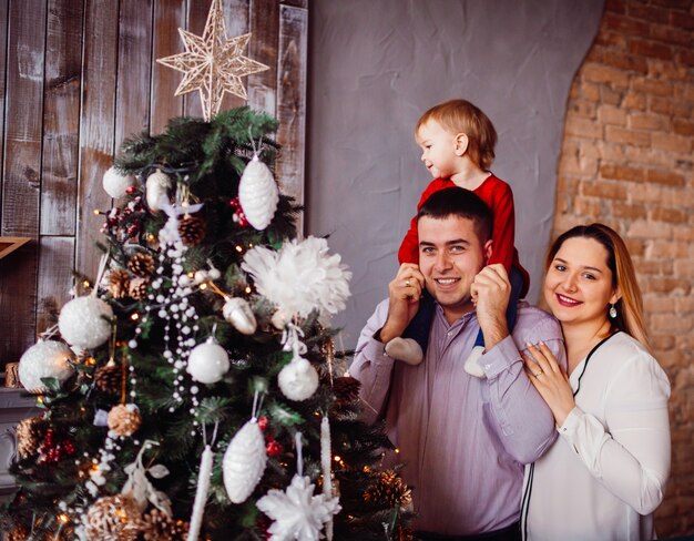 Papa tient la petite fille sur son cou posant avec maman devant un arbre de Noël riche