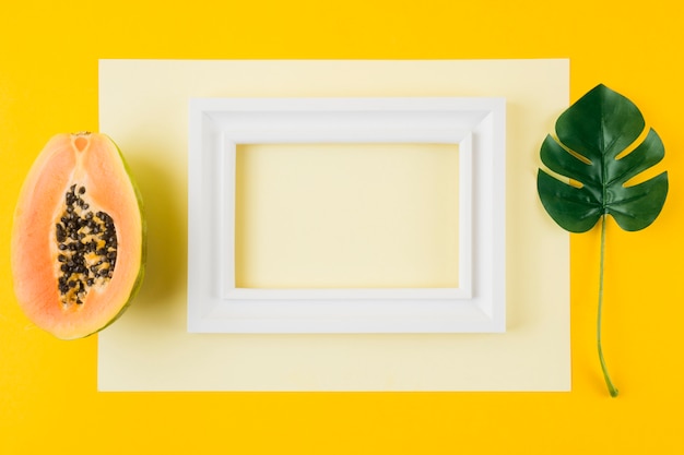 Photo gratuite papaye coupée en deux; feuille de monstera et cadre en bois blanc sur papier sur fond jaune