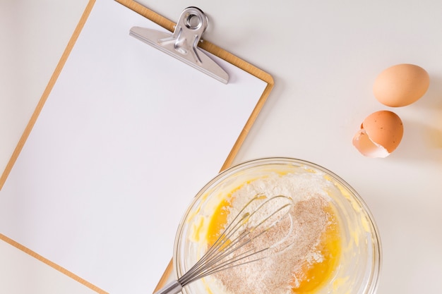 Papier blanc blanc sur le presse-papiers avec oeuf fouetté et bol de farine sur fond blanc