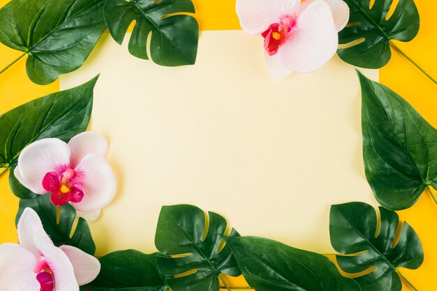 Papier blanc entouré de feuilles artificielles et de fleurs d&#39;orchidées sur fond jaune