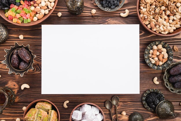 Photo gratuite papier blanc vierge entouré d'un mélange de fruits secs; rendez-vous; lukum; baklava et noix sur un bureau en bois pour ramadan