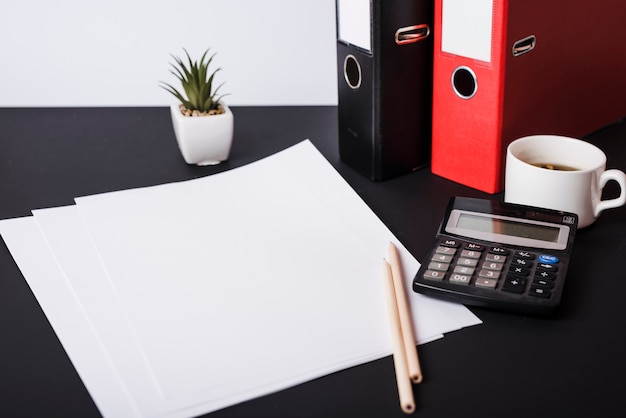 Papiers vierges blancs; des crayons; plante en pot; dossiers papier; tasse à café et calculatrice sur bureau noir