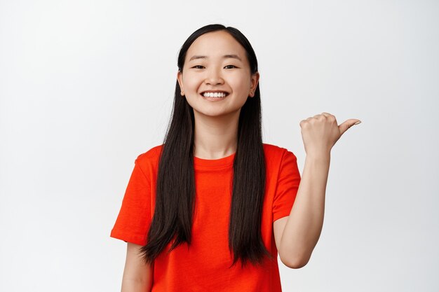 Par ici. Femme asiatique souriante démontrant la bannière, montrant le chemin, pointant le doigt vers la droite avec un sourire satisfait, debout sur blanc