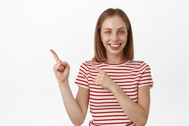 Par ici. Une fille blonde souriante et amicale pointant les doigts vers la gauche et regardant la caméra avec plaisir, montrant une publicité de réduction de vente, pointant vers la bannière, fond blanc.