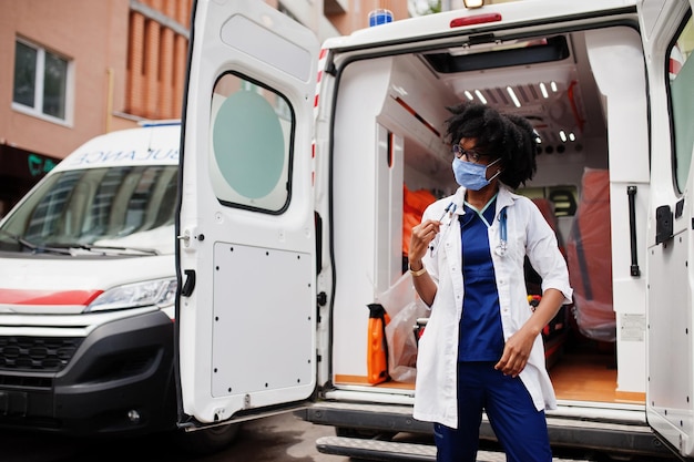 Paramédic féminin afro-américain dans le masque médical de protection du visage debout devant la voiture d'ambulance