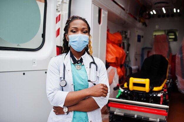 Paramédic féminin afro-américain dans le masque médical de protection du visage debout devant la voiture d'ambulance