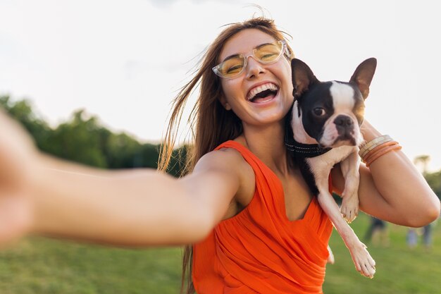 Parc de jolie femme heureuse faisant selfie photo, tenant un chien boston terrier, souriant humeur positive, style estival branché, vêtu d'une robe orange, lunettes de soleil, jouer avec un animal de compagnie, s'amuser
