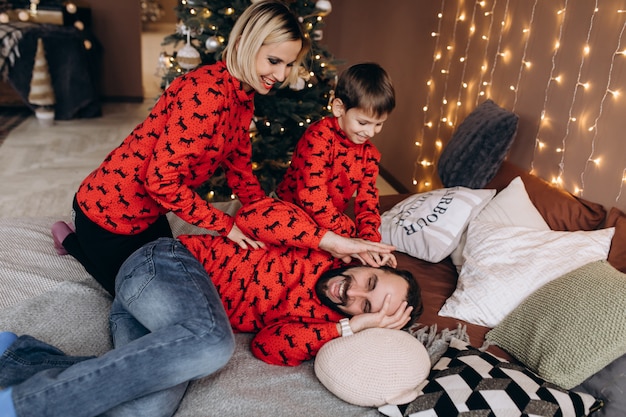 Des parents attirants et leur petit fils en pull rouge s’amusent sur le lit avant un Noël