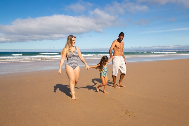 Parents et petite fille portant des maillots de bain, marchant sur le sable doré de la mer