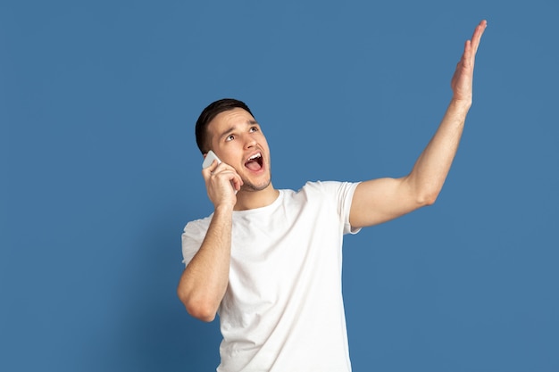 Parler au téléphone, pointer du doigt. Portrait de jeune homme caucasien sur fond bleu studio. Beau modèle masculin dans un style décontracté, couleurs pastel. Concept d'émotions humaines, expression faciale, ventes.