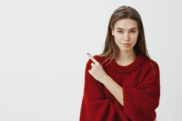 Partager un endroit secret avec nous. Confiante belle femme entrepreneur en pull ample rouge, souriant avec une expression assurée intriguée, pointant dans le coin supérieur gauche