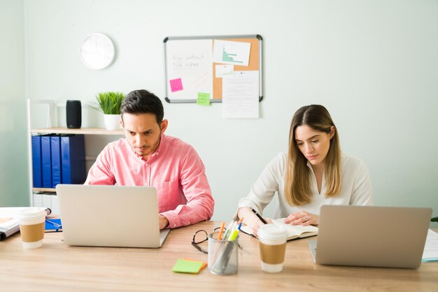 Partenaires commerciaux professionnels travaillant côte à côte à un bureau. Femme caucasienne écrivant sa tâche sur un agenda et collègue masculin tapant sur un ordinateur portable